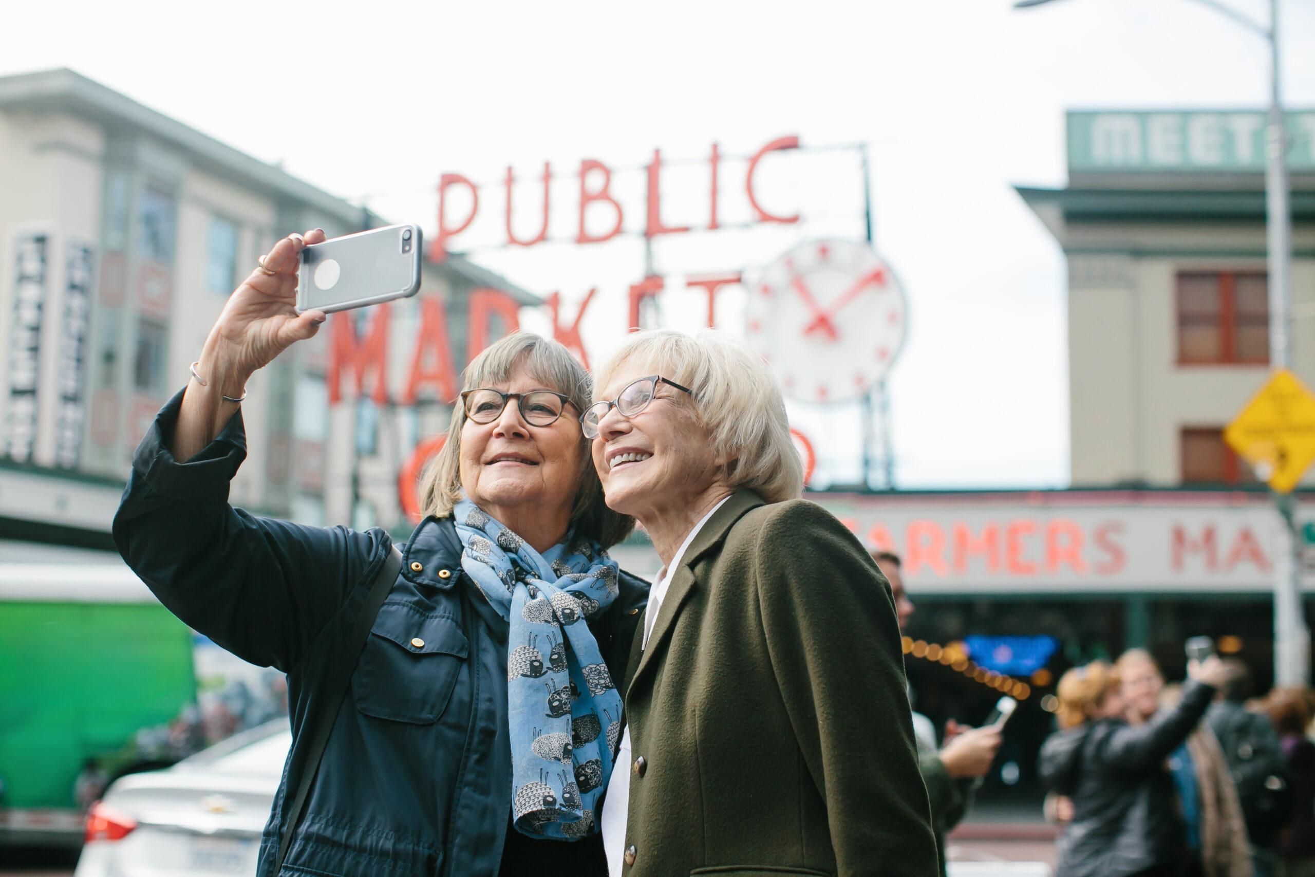 Two women take a selfie in front of a Public Market neon sign.